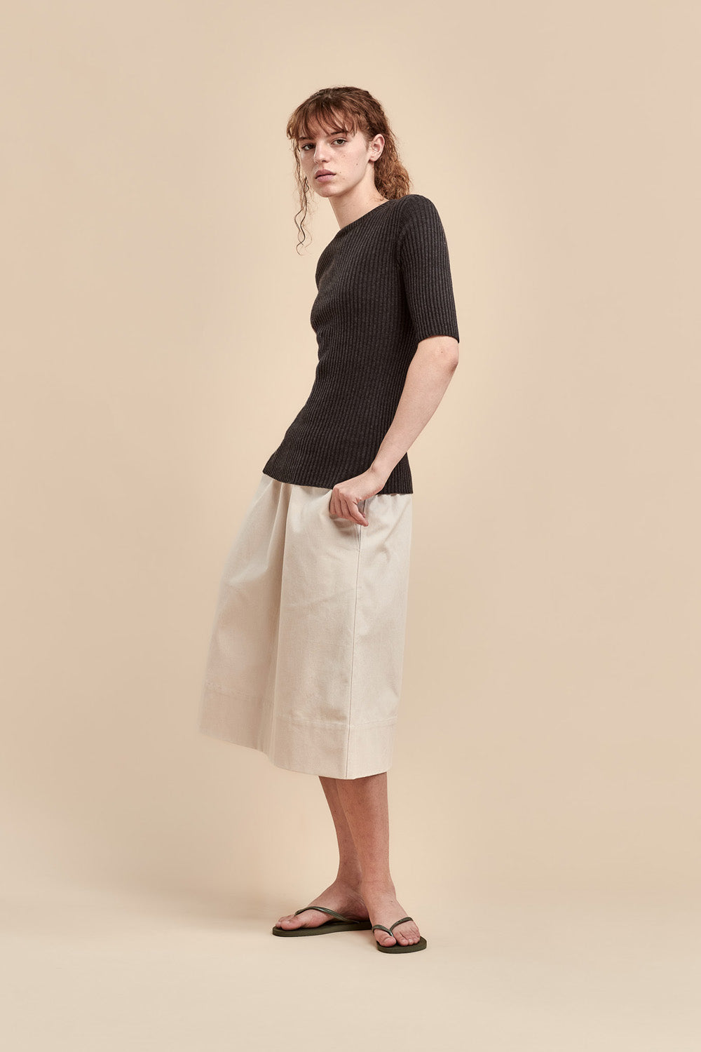 A woman modeling a dark short-sleeve ribbed knit top and a light beige midi skirt, paired with black flip-flops, against a solid beige studio background.