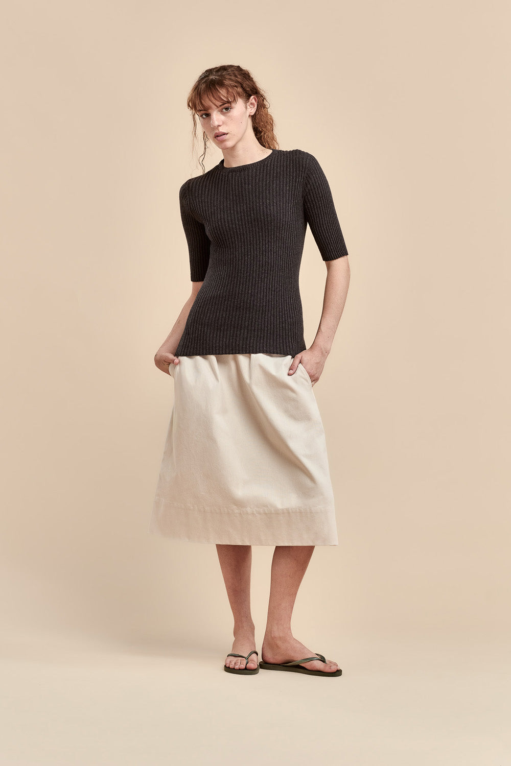 A woman modeling a dark ribbed short-sleeve top and a cream-colored skirt against a solid beige studio background.