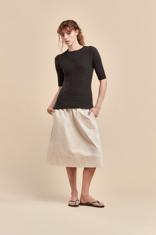 A woman modeling a dark ribbed short-sleeve top and a cream-colored skirt against a solid beige studio background.