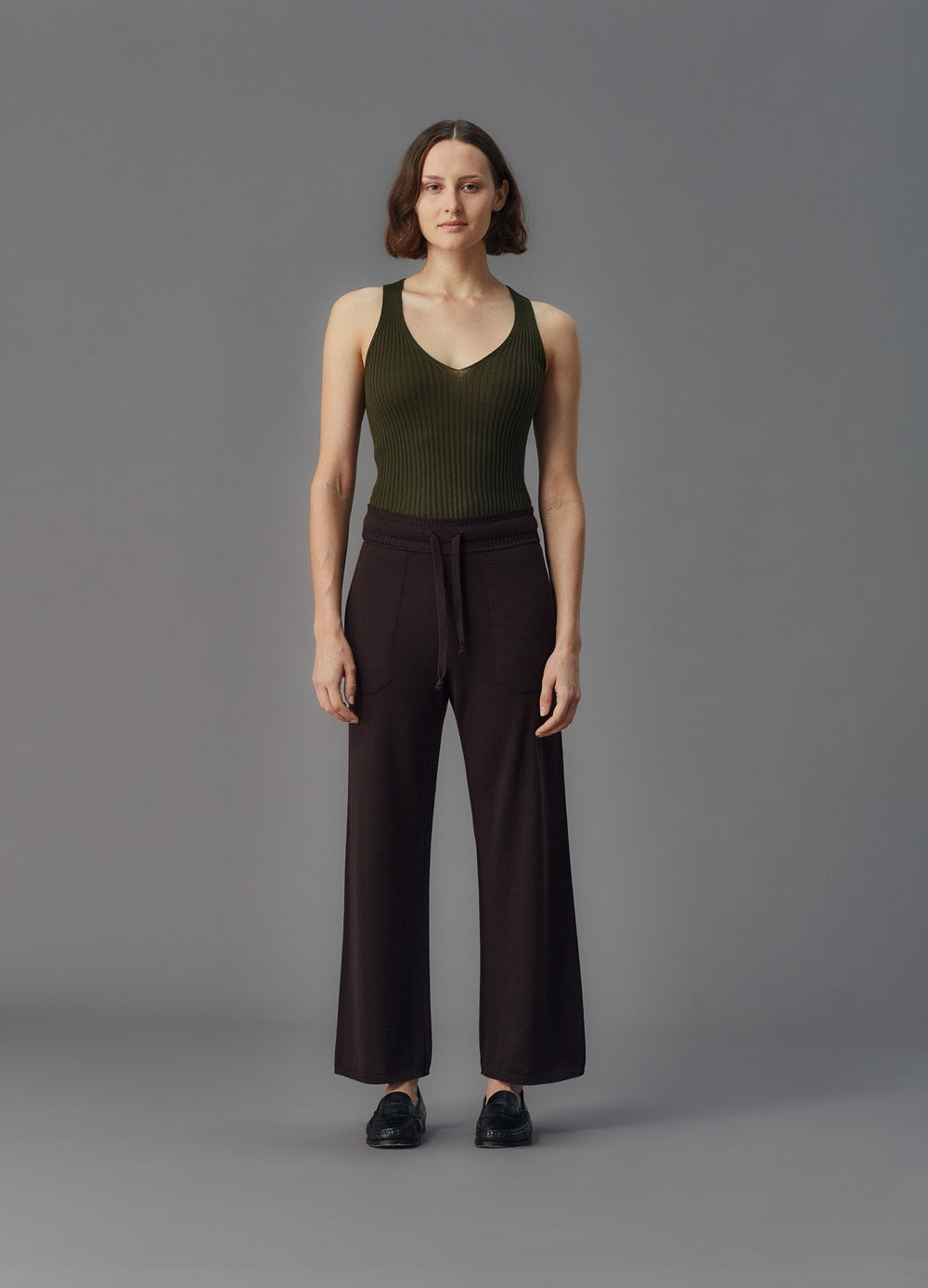 Woman wearing a green tank top and brown pants against a gray background