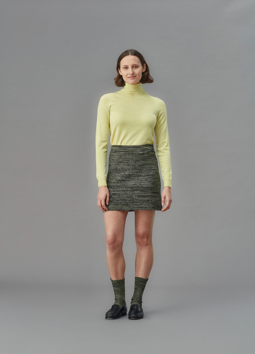 Woman wearing a light yellow skivvy turtleneck sweater and knit skirt against a gray background