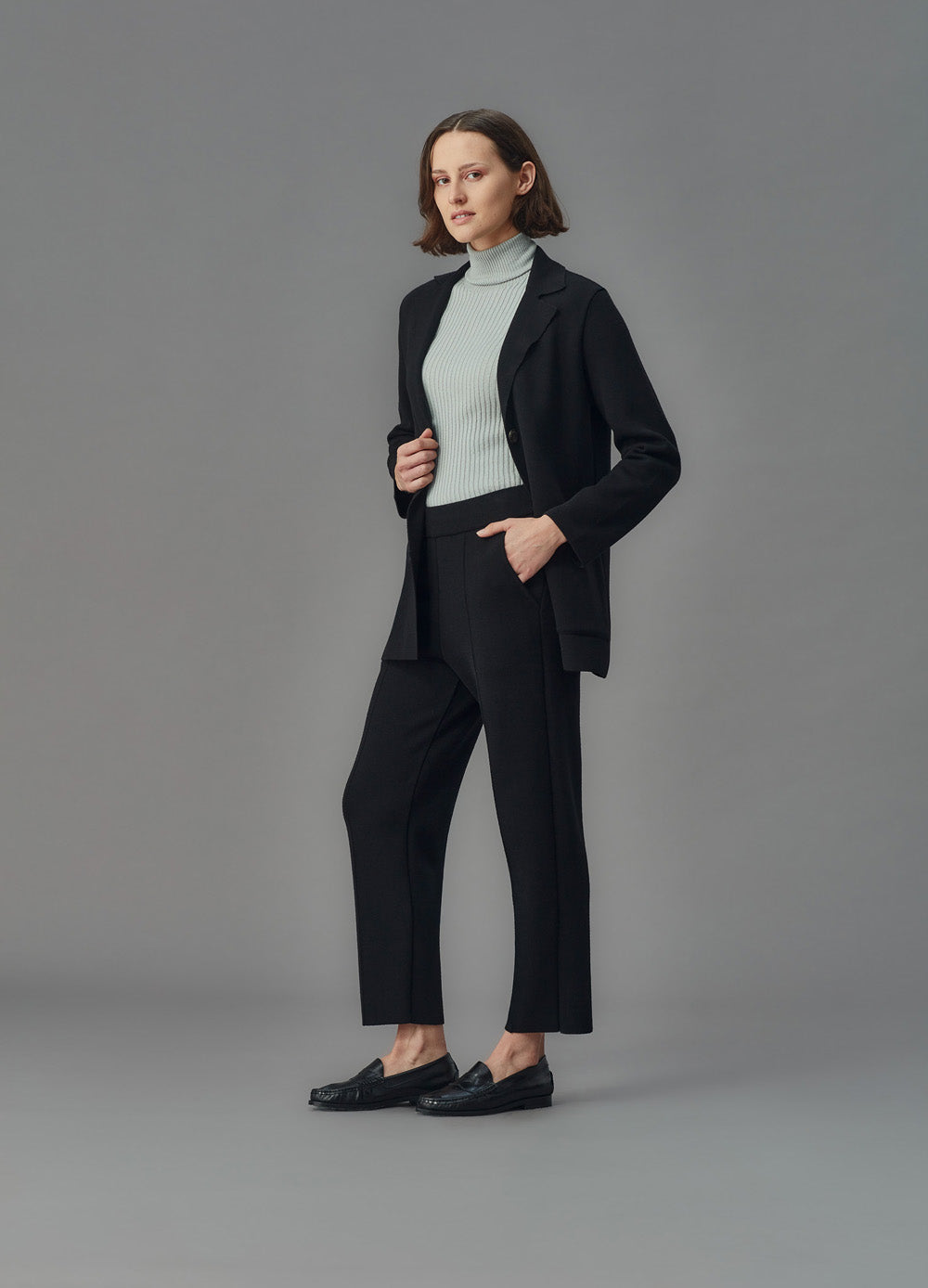 Woman wearing a black merino jacket and pants with a light blue turtleneck against a gray background