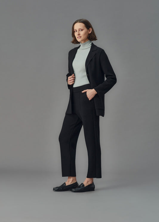Woman wearing a black merino jacket and pants with a light blue turtleneck against a gray background