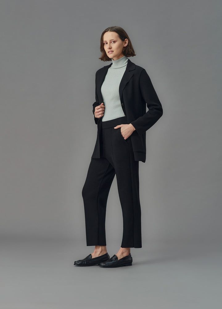 Woman wearing a black merino jacket and pants with a light blue turtleneck against a gray background