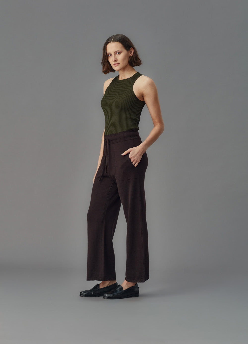 Woman wearing a green ribbed merino tank top and dark pants against a gray background