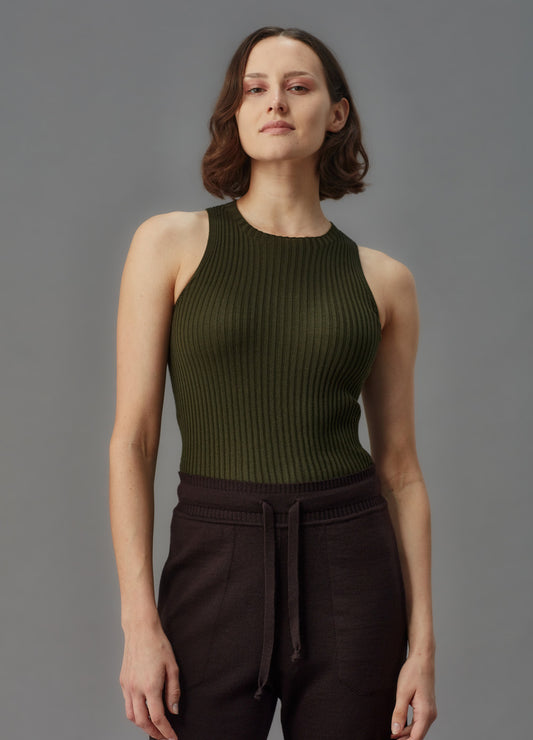 Woman wearing a green ribbed merino tank top and dark pants against a gray background
