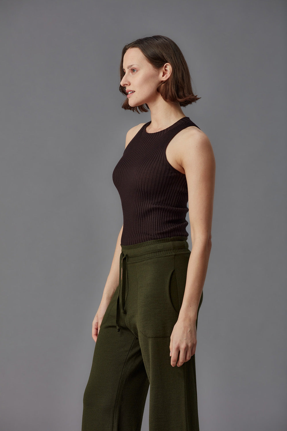 Woman wearing a brown ribbed merino tank top and green pants against a gray background