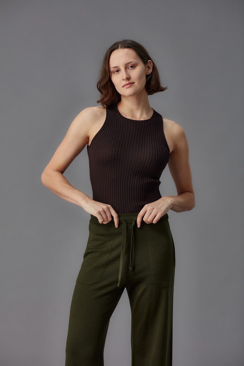Woman wearing a brown ribbed merino tank top and green pants against a gray background