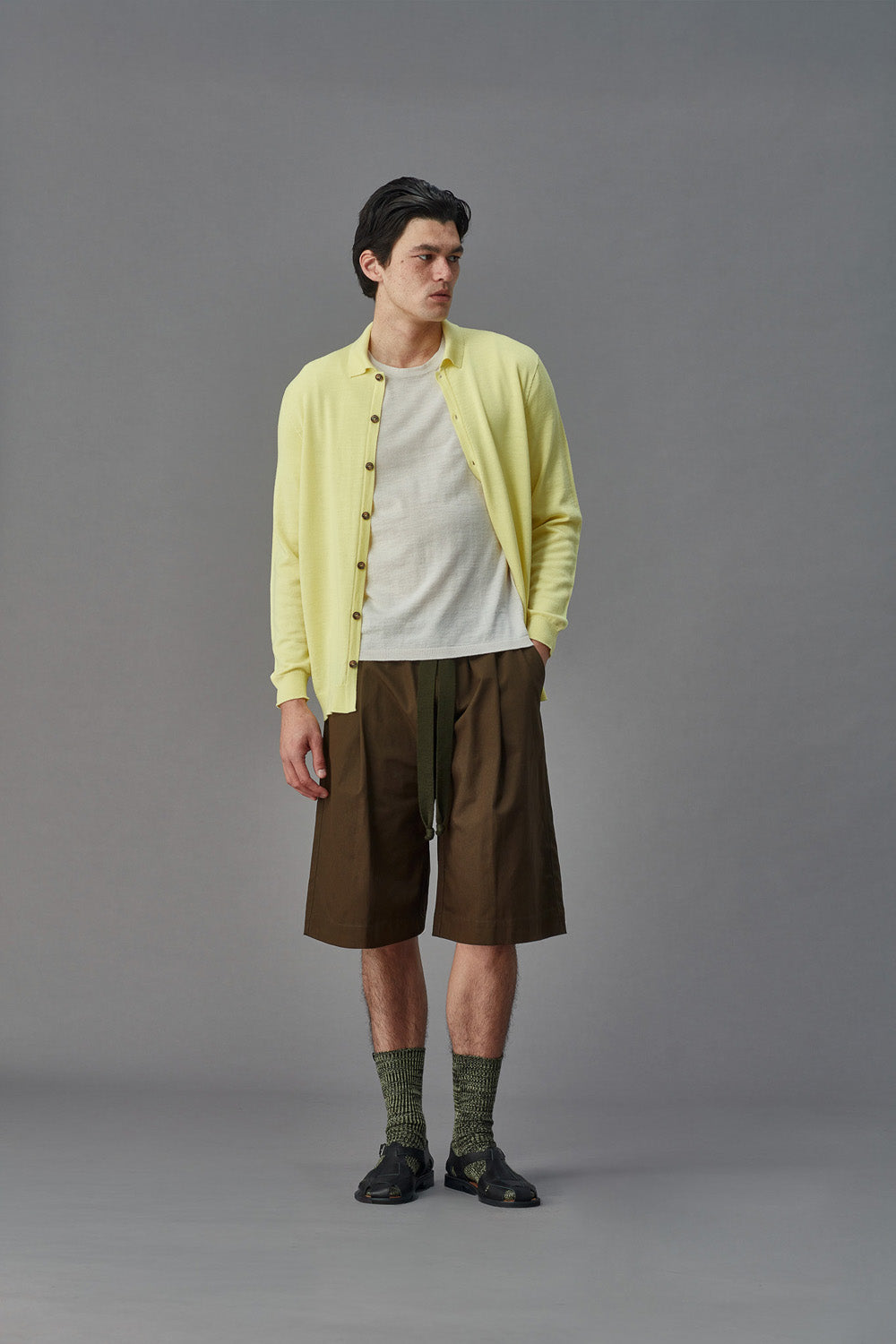 Man wearing a light yellow cardigan over a white shirt with brown shorts on a gray background