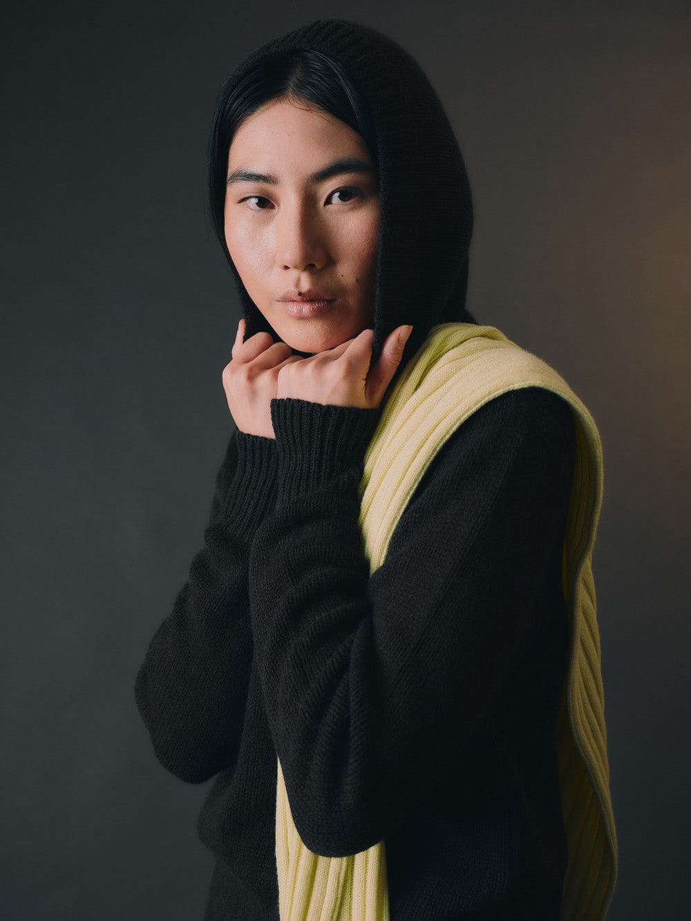 Person wearing a black hooded knit sweatshirt and yellow scarf against a dark background