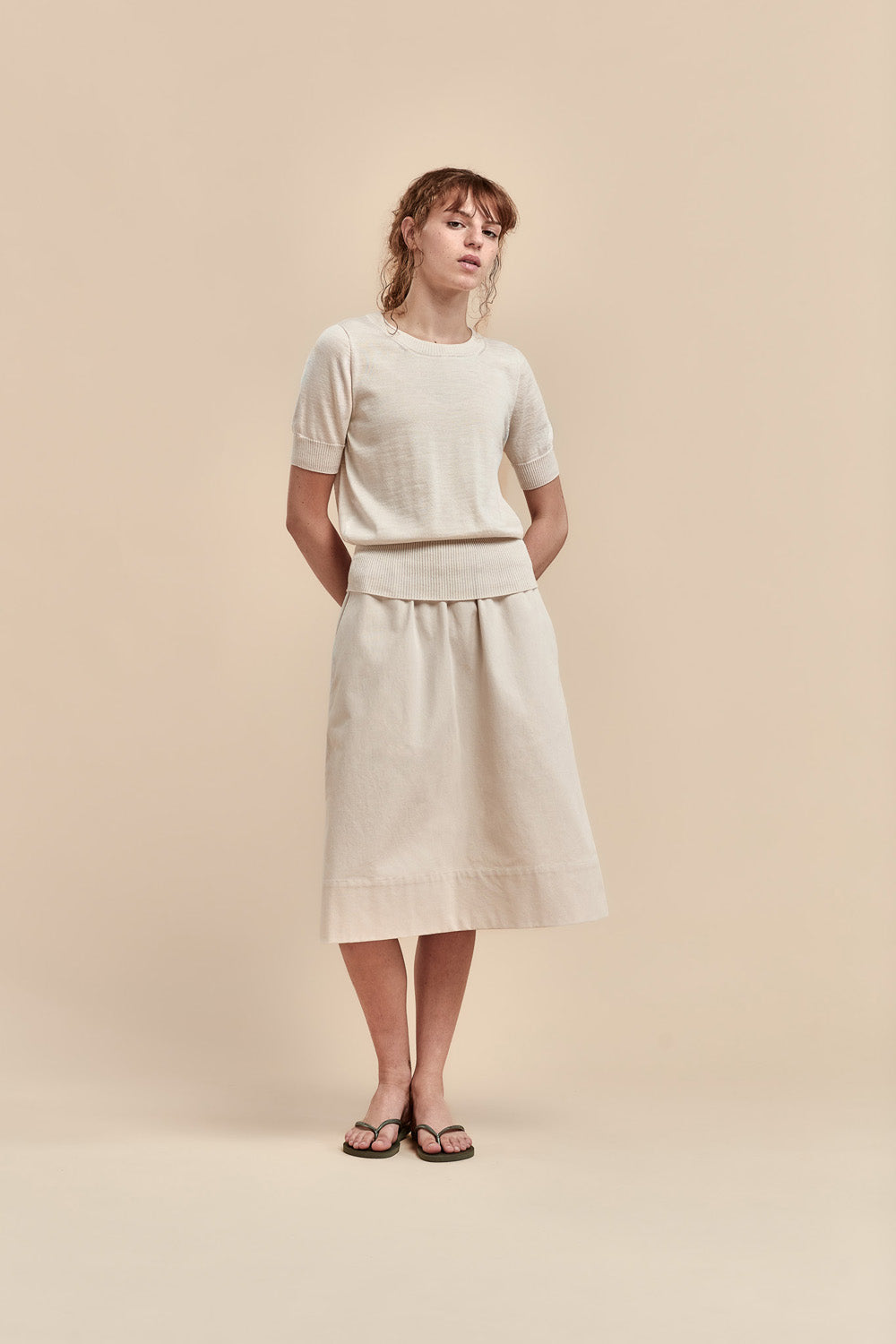 A woman modeling a light merino short-sleeve crewneck top and a matching midi skirt, paired with black sandals, against a solid beige studio background.