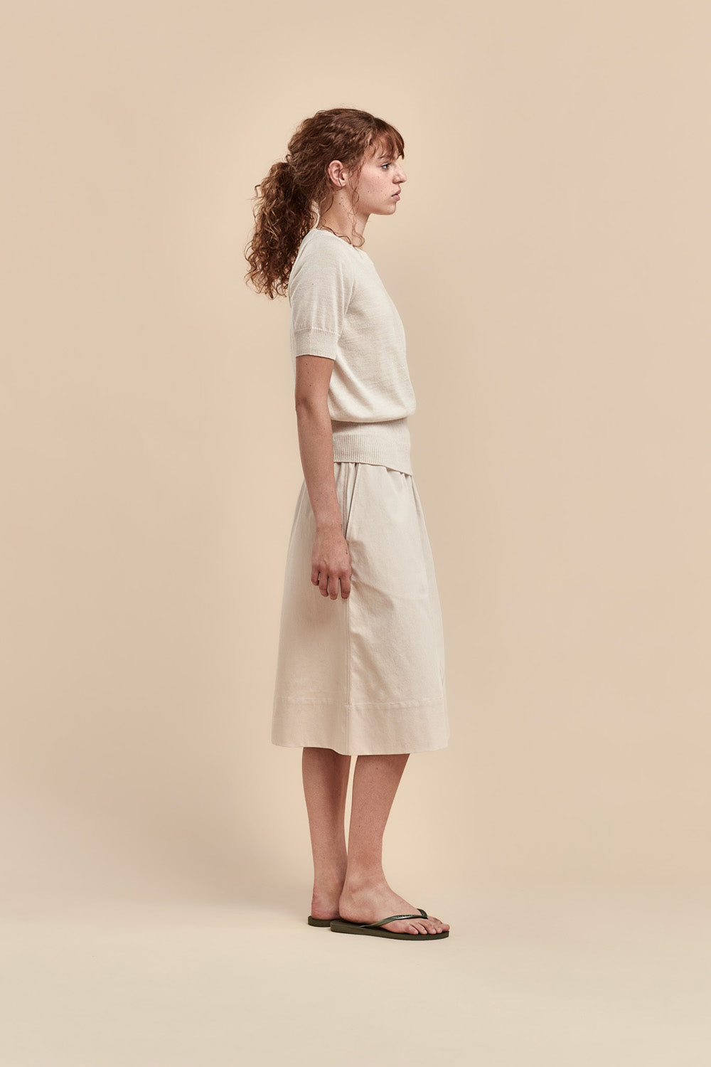 A woman stands in profile wearing a light merino short-sleeve top and matching midi skirt, paired with dark flip-flop sandals, against a solid beige studio background.