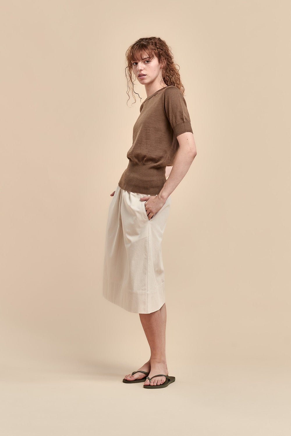 A woman stands in profile wearing a brown short-sleeve merino knit top, midi skirt with side pockets, and dark sandals against a solid beige studio background.