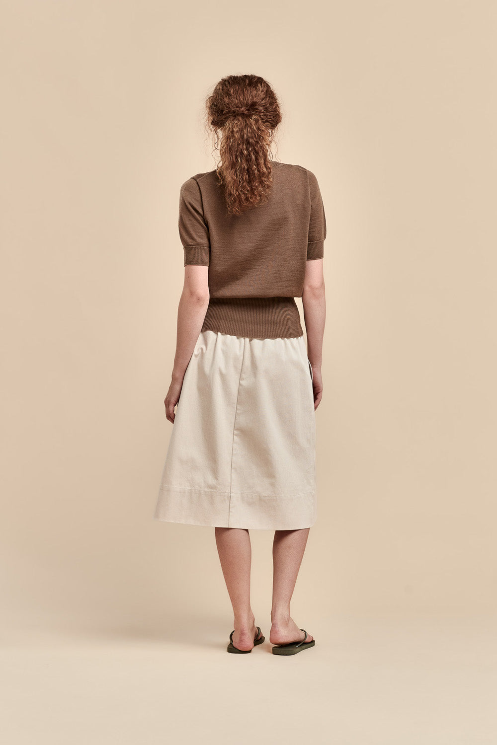 A woman is shown from behind wearing a brown merino short-sleeve knit top and a cream-colored midi skirt, standing against a solid beige studio background.
