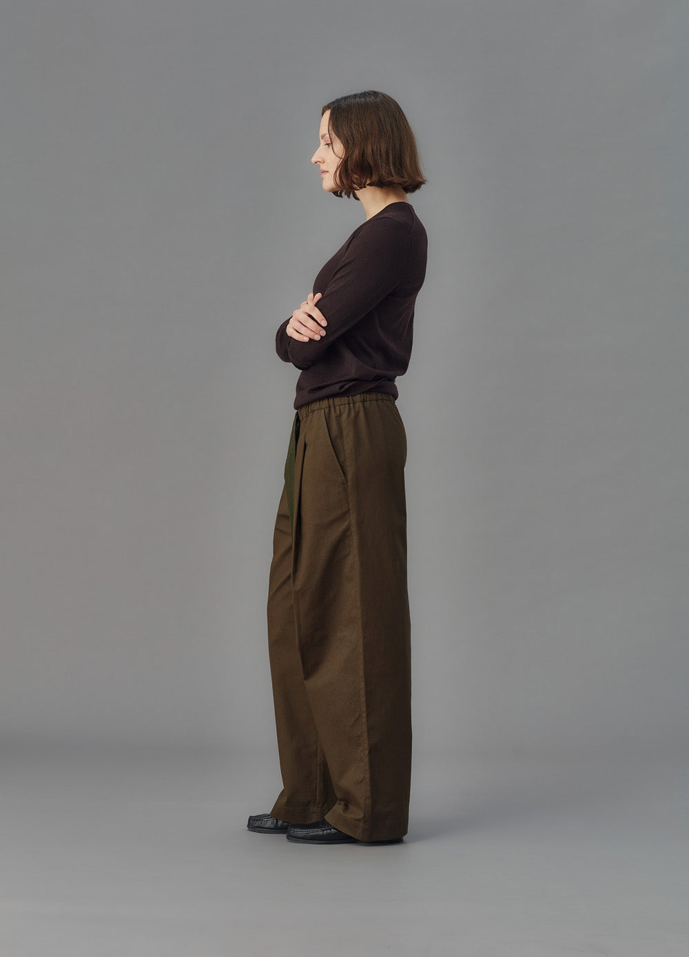 Person wearing a dark sweater and green pants standing against a gray background