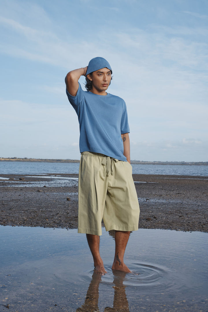 Man standing on a beach wearing a blue hat and shirt and beige shorts, with a clear sky in the background.