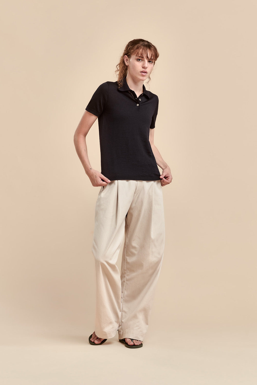 A woman modeling a black short-sleeve merino polo shirt and cream wide-leg trousers against a solid beige studio background.