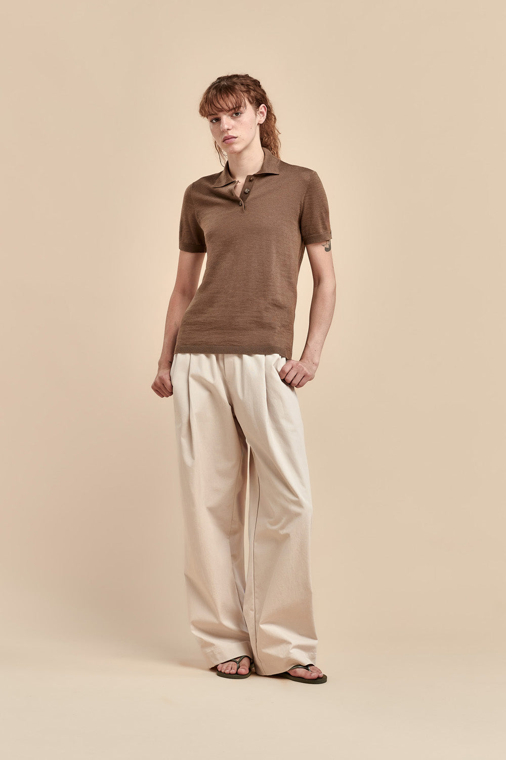 A woman modeling a brown merino short-sleeve polo shirt with wide-leg cream trousers and black sandals, standing against a solid beige studio background.