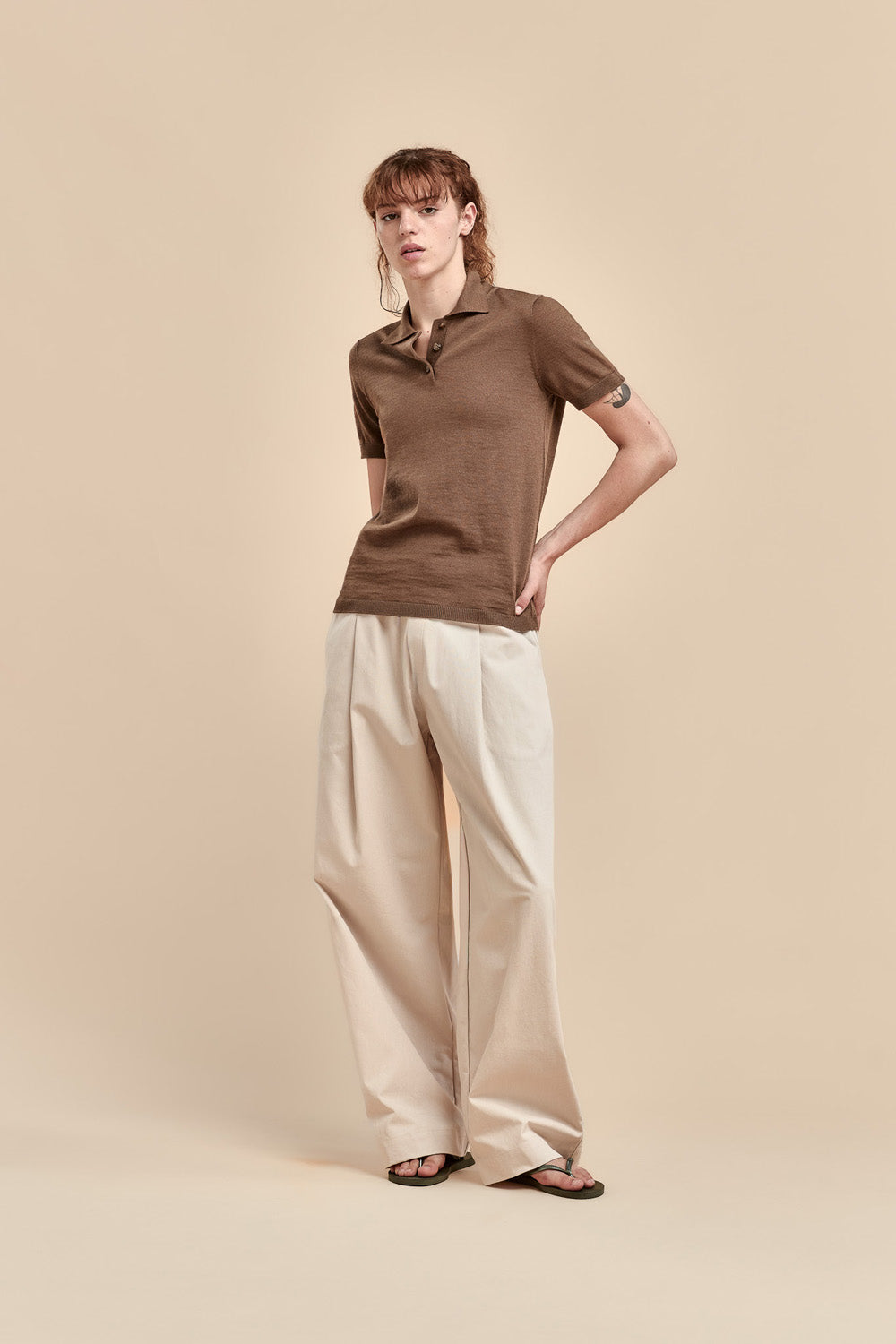 A woman modeling a brown merino short-sleeve polo shirt with wide-leg cream trousers and black sandals, standing against a solid beige studio background.
