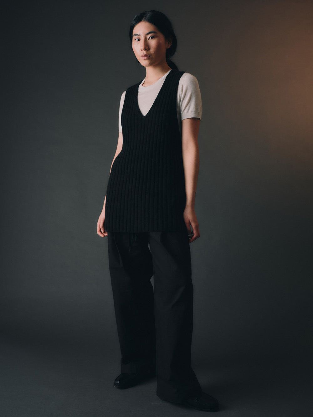 Woman wearing a black knitted vest over a white shirt against a dark background.