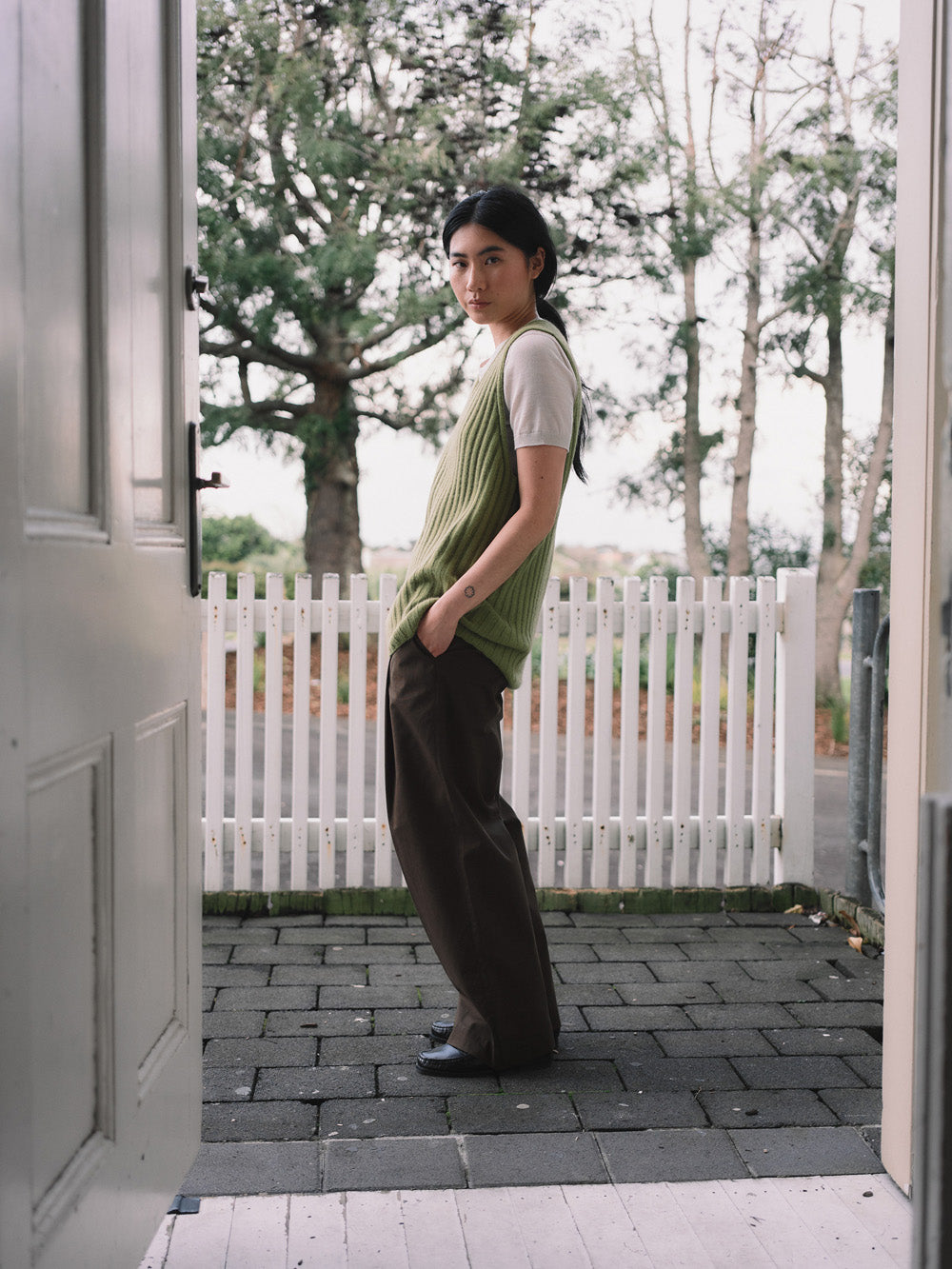 Woman standing in a doorway wearing a green knitted vest and dark pants, with a white picket fence and trees in the background.