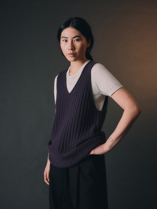 Woman wearing a dark purple knitted vest over a white shirt against a dark background.