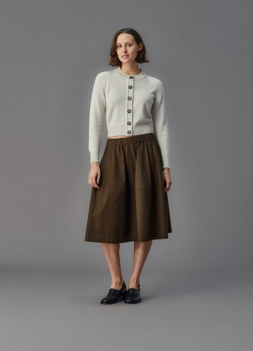 Woman wearing a alabaster cardigan and brown skirt against a gray background