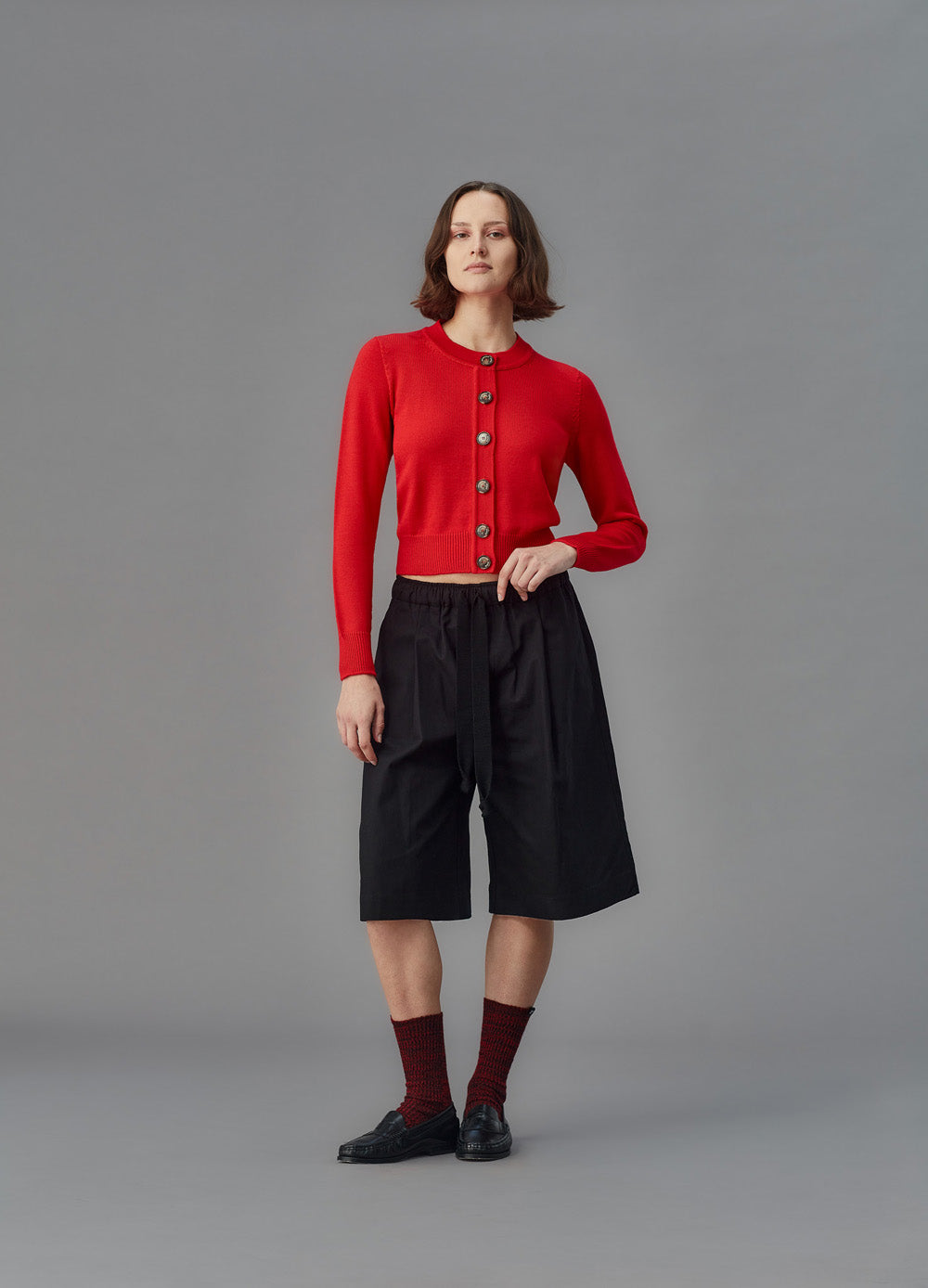 Woman wearing a red cardigan, black shorts, and black shoes on a gray background