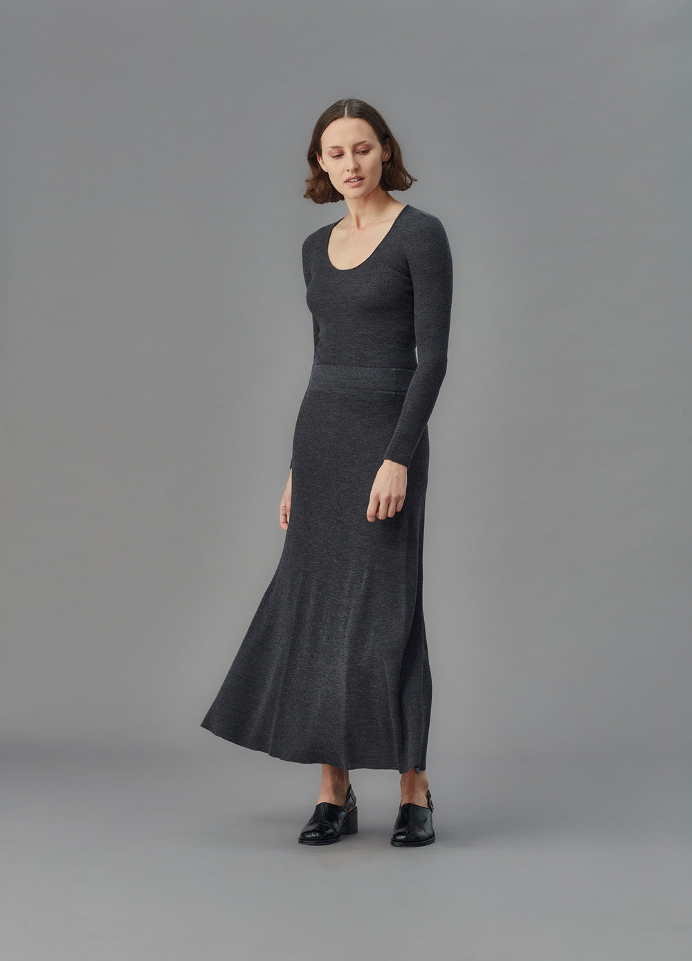 Woman wearing a long dark gray u top and skirt against a gray background