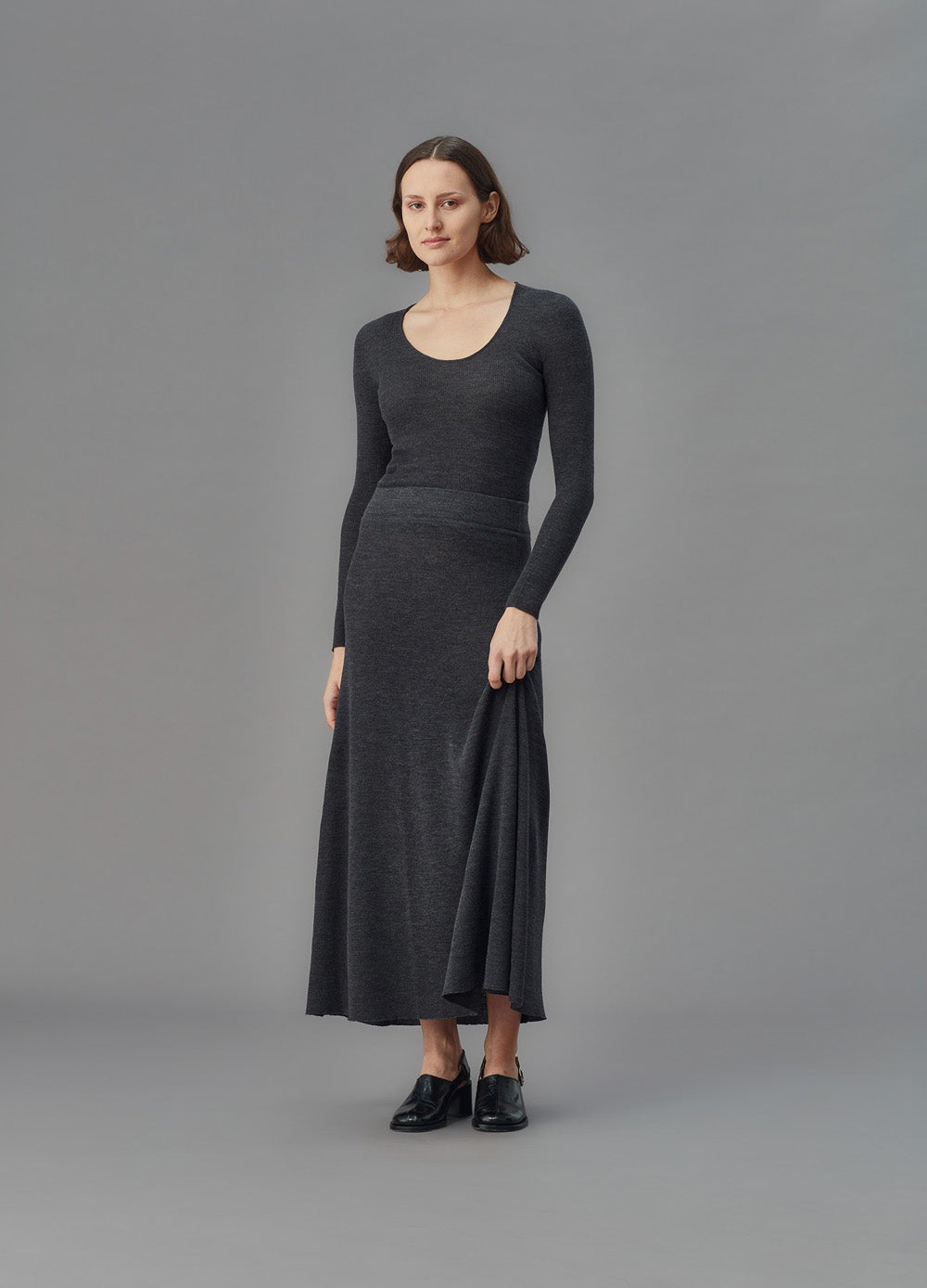 Woman wearing a long dark gray u top and skirt against a gray background