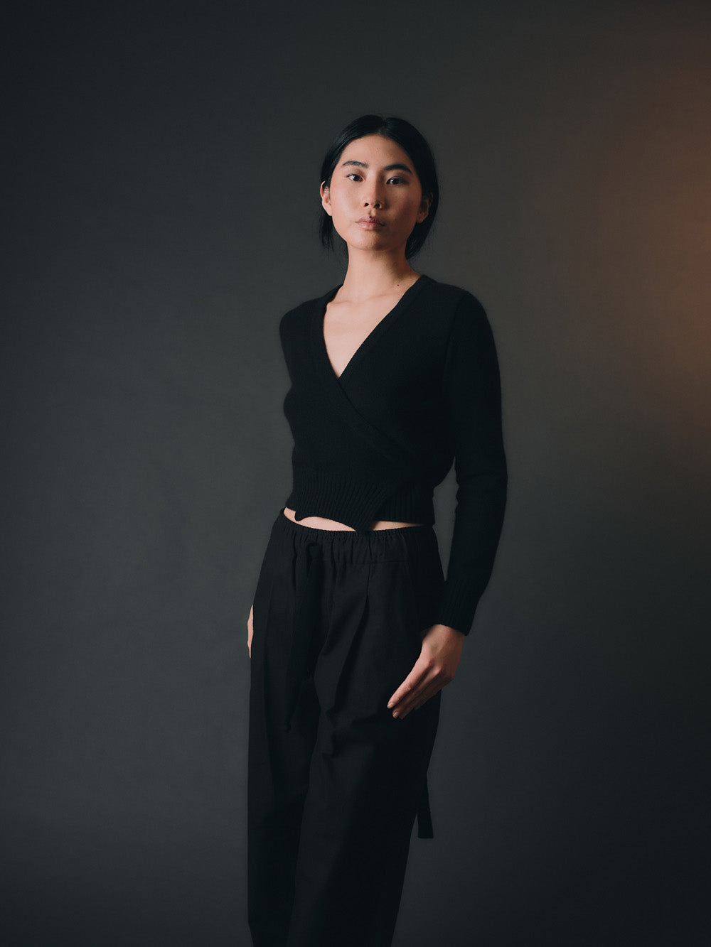 Woman wearing a black knitted wrap sweater and black wide-leg pants against a dark background