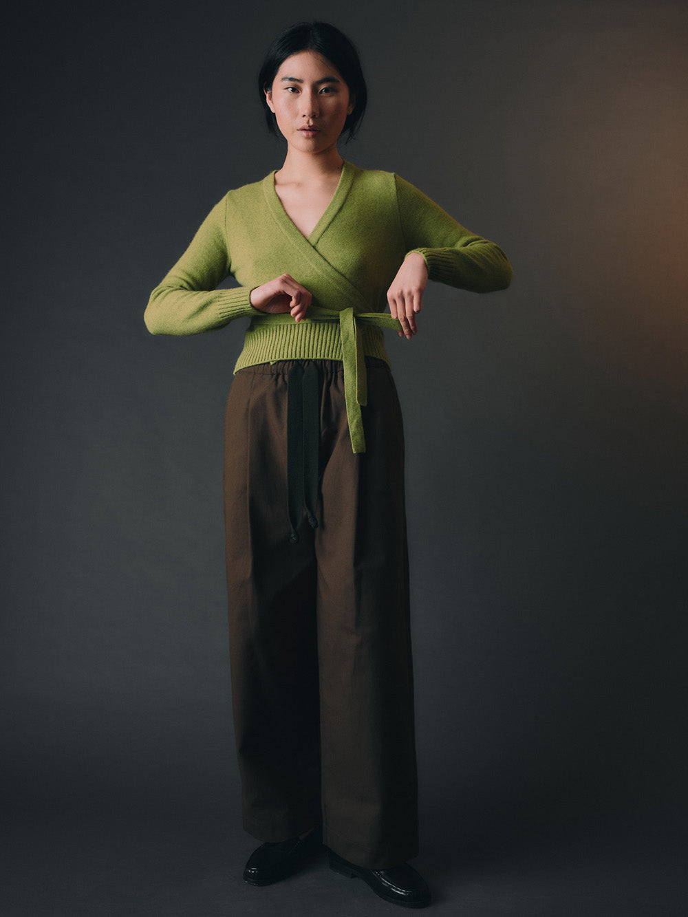 Woman wearing a green knitted wrap sweater and brown wide-leg pants against a dark background