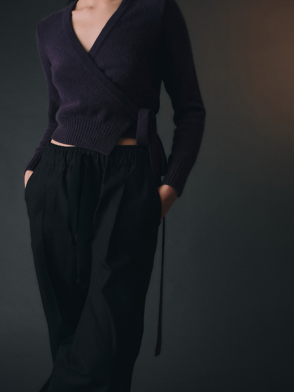 Woman wearing a purple knitted wrap sweater and black wide-leg pants against a dark background
