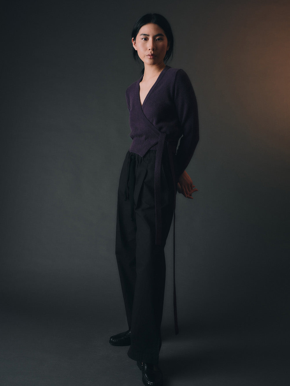 Woman wearing a purple knitted wrap sweater and black wide-leg pants against a dark background
