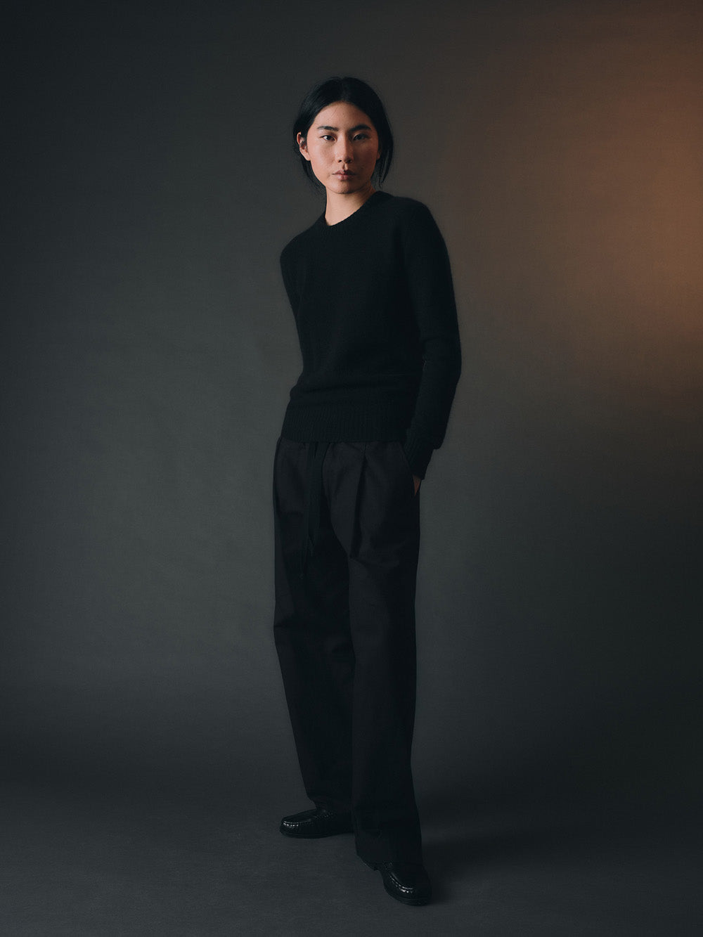 Person wearing a black sweater and trousers against a dark background