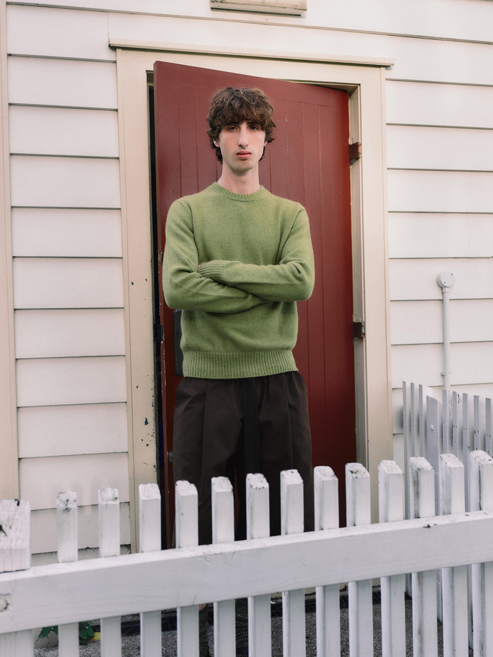 Person wearing a green sweater standing in front of a red door with a white picket fence.