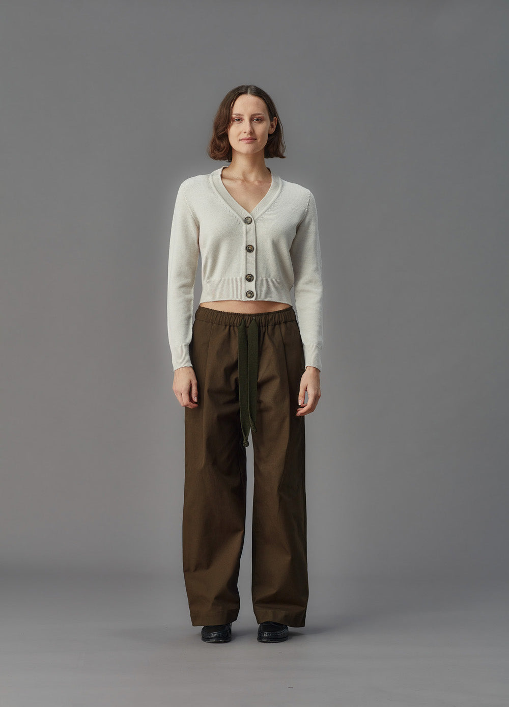 Woman wearing a alabaster white merino cardigan and brown pants on a gray background