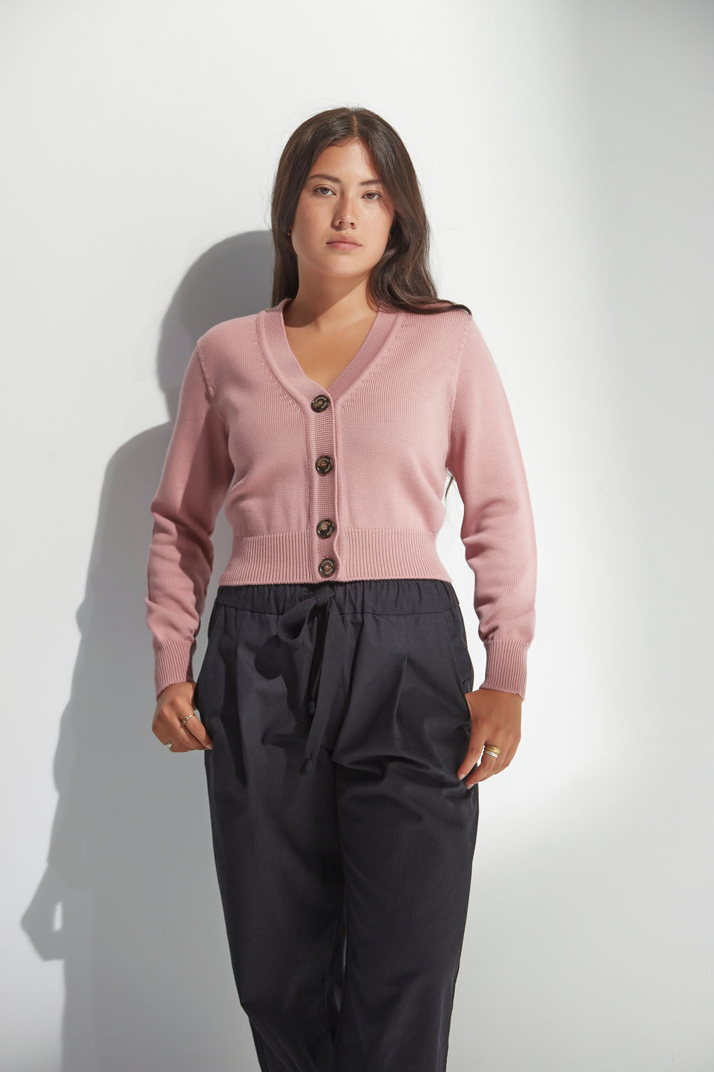 Woman wearing a pink v neck cardigan and black pants against a white background