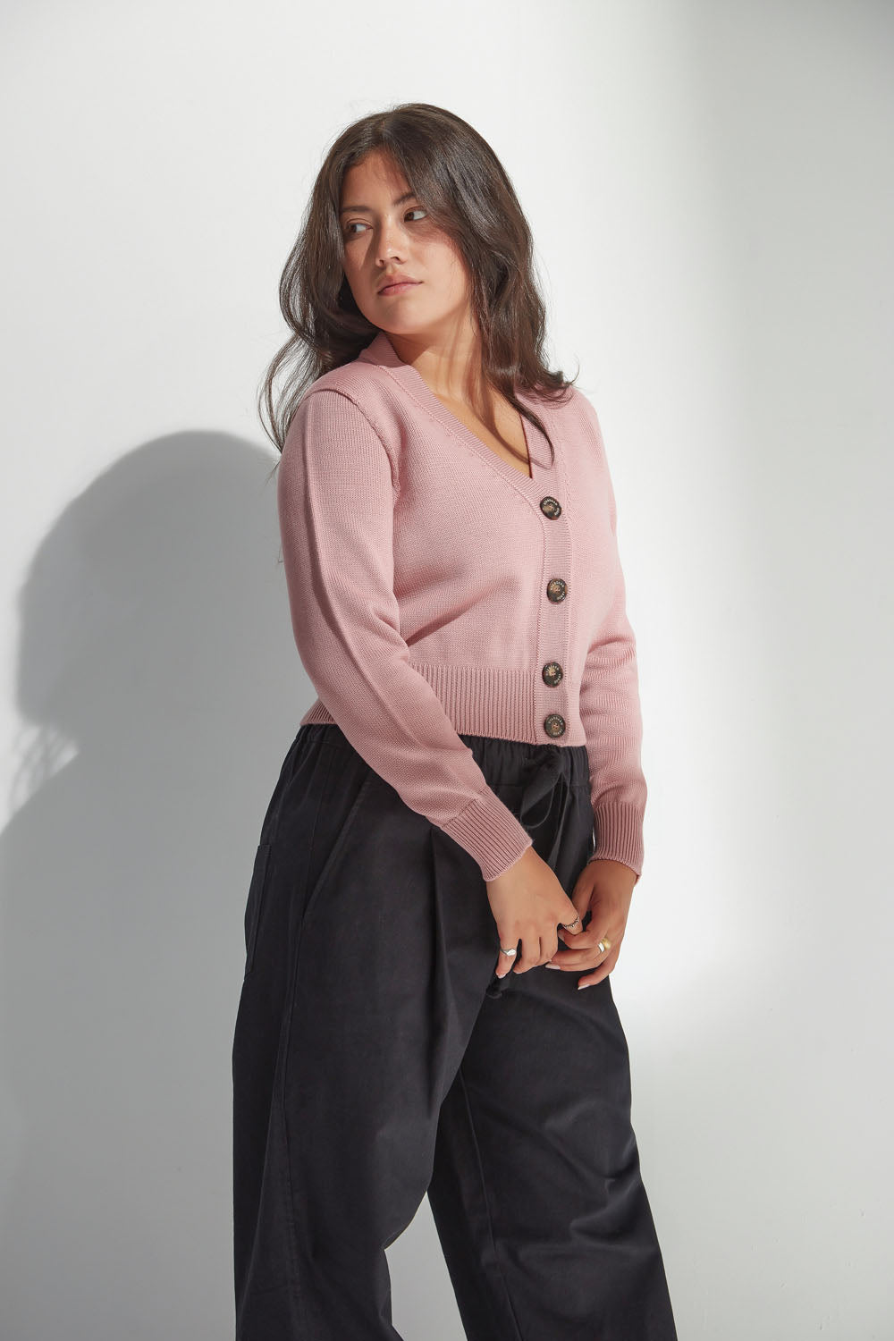 Woman wearing a pink v neck cardigan and black pants against a white background