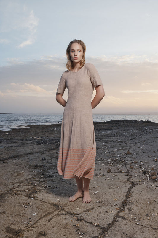 Woman in a beige dress standing on a rocky beach with ocean and sky in the background.