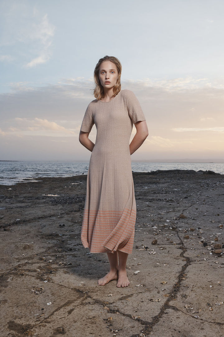 Woman in a beige dress standing on a rocky beach with ocean and sky in the background.