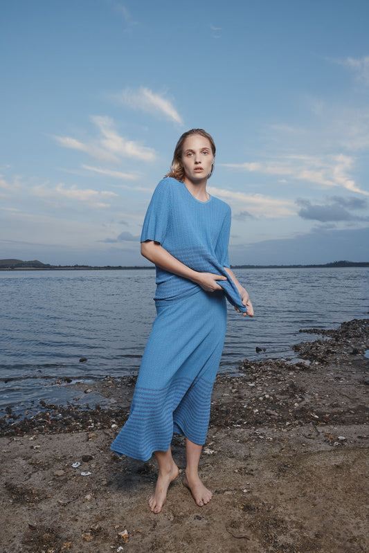 Woman in a blue skirt and top standing on a beach with a body of water and sky in the background.