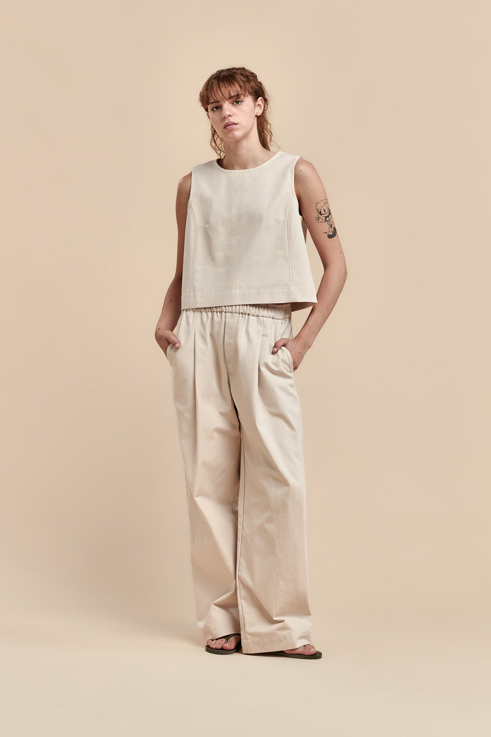 A woman modeling a light beige two-piece set consisting of a sleeveless cropped top and matching wide-leg trousers, standing against a solid beige studio background.