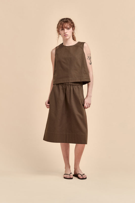 A model stands against a solid beige studio background wearing a sleeveless, cropped brown top and a matching brown midi skirt, paired with black flip-flop sandals.