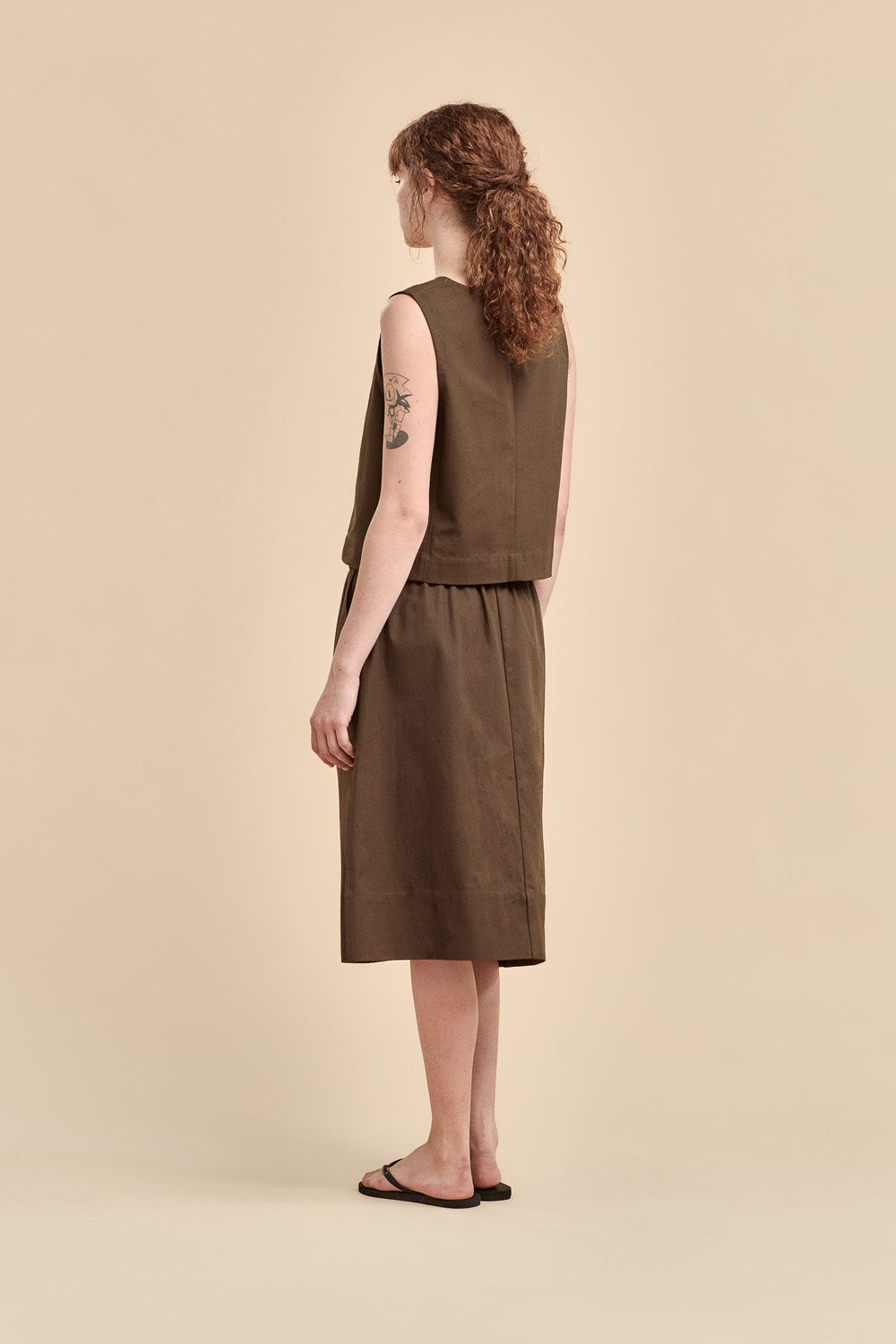 A woman with curly brown hair is shown from the back wearing a sleeveless cotton broadcloth top and matching midi skirt set against a solid beige studio background.