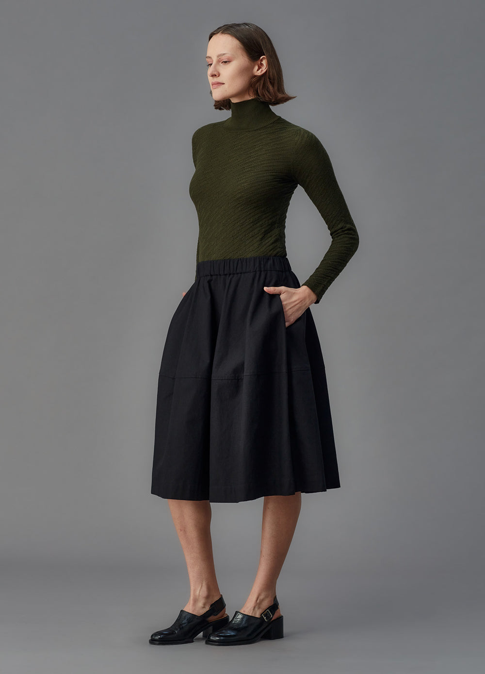 Woman wearing a green turtleneck and black skirt against a gray background