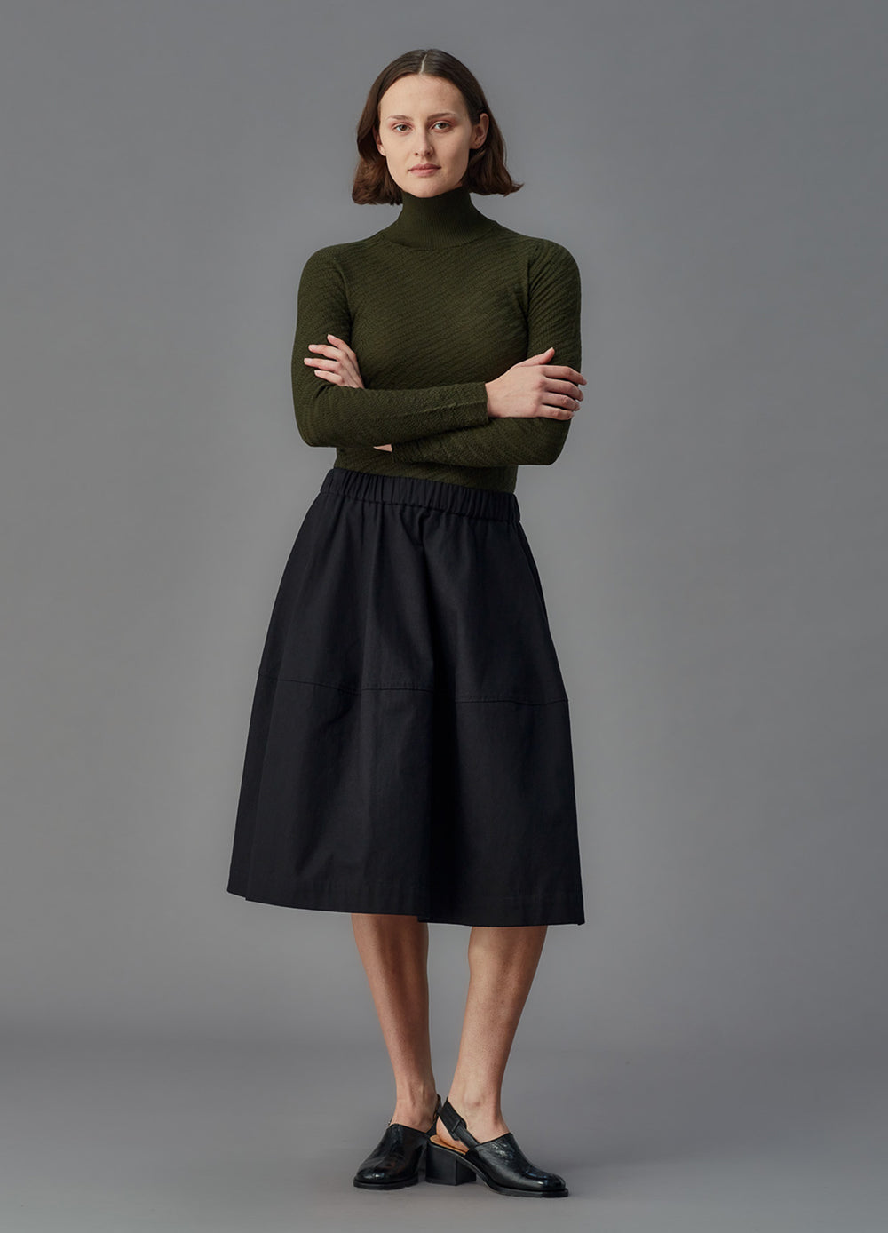 Woman wearing a green turtleneck and black skirt against a gray background
