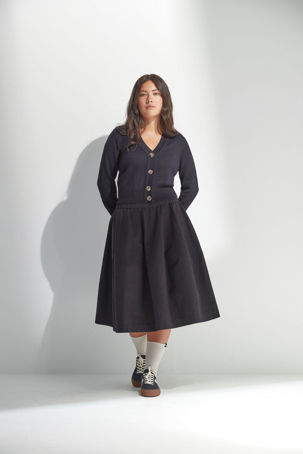 Woman wearing a black skirt and buttoned cardigan standing against a white wall.