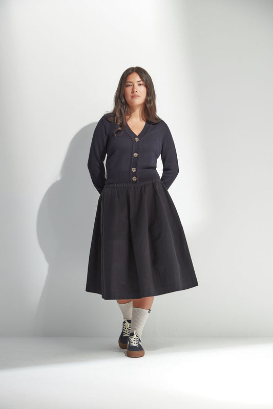 Woman wearing a black skirt and buttoned cardigan standing against a white wall.