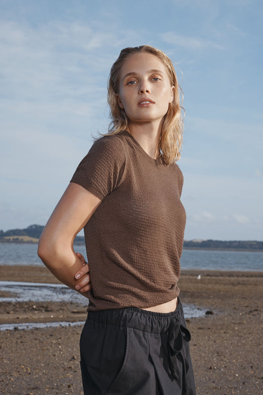 Woman standing on a beach wearing a brown t-shirt and black pants with a blue sky in the background.